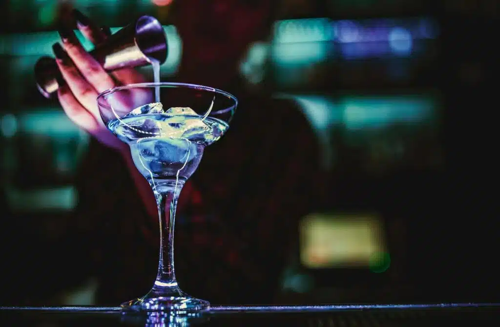 A bartender in a dimly lit bar pours a drink from a metal jigger into a martini glass filled with ice.