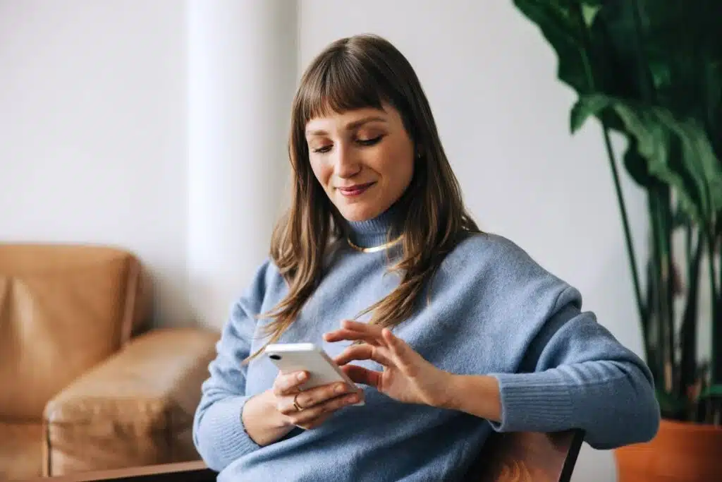 A woman in a blue sweater taking an online course on her phone while sitting in a chair.